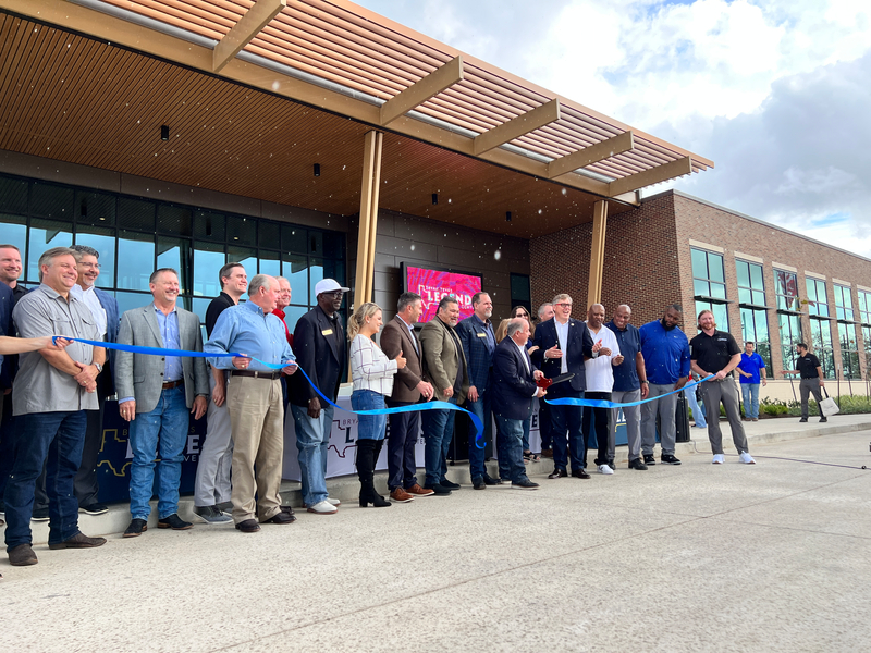 A diverse group of people stand smiling in front of a modern building, cutting a blue ribbon at a grand opening event, conveying excitement and celebration.